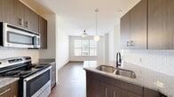 This efficiently designed single-floor unit offers a thoughtful layout across four distinct spaces: a living room, kitchen, bathroom, and closet. The open-concept kitchen flows seamlessly into the living area, creating a cohesive and functional living space. The kitchen is appointed with rich dark wood cabinetry, granite-look countertops, a double stainless steel sink, and a full suite of stainless steel appliances including a refrigerator, electric range, microwave, and dishwasher. A white subway tile backsplash adds a clean, modern touch. A pendant light fixture and ceiling fan with light illuminate the transition between the kitchen and living area, enhancing the airy ambiance. The living room benefits from multiple windows dressed with horizontal blinds, allowing abundant natural light to fill the space. Hardwood-style flooring runs throughout the unit, providing warmth and visual continuity. The bathroom features a combined tub and shower with white tile surround, a granite-topped vanity with dark cabinetry, and oil-rubbed bronze fixtures. Crisp white walls and clean lines throughout give the unit a bright, contemporary feel well-suited for modern living.