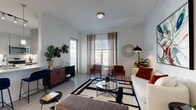 This well-appointed single-floor residence combines contemporary design with functional living across a thoughtfully arranged floor plan. The open-concept kitchen is a standout feature, outfitted with stainless steel appliances including a refrigerator, electric range, and over-the-range microwave, complemented by sleek grey cabinetry, quartz countertops, and a peninsula with bar seating for two. Pendant lighting and track fixtures illuminate the space, adding to its modern aesthetic. Light wood-style flooring flows throughout, creating a cohesive look across all areas. The living room offers generous space for relaxation, with large windows dressed in sheer curtains that invite natural light. Adjacent to the entryway, a compact dining area accommodates an intimate table setting beneath framed artwork, establishing a welcoming atmosphere. The bedroom is a serene retreat, featuring windows with outdoor views, ample room for furnishings, and soft natural light filtered through blinds and curtains. The bathroom is clean and modern, with a wide vanity featuring quartz countertops, a vessel sink, brushed chrome fixtures, a large mirror, and a tiled tub-shower combination. Throughout the residence, white walls and light flooring create a bright, airy ambiance.