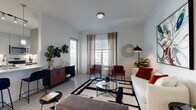 This well-appointed single-floor residence combines contemporary design with functional living across a thoughtfully arranged floor plan. The open-concept kitchen is a standout feature, outfitted with stainless steel appliances including a refrigerator, electric range, and over-the-range microwave, complemented by sleek grey cabinetry, quartz countertops, and a peninsula with bar seating for two. Pendant lighting and track fixtures illuminate the space, adding to its modern aesthetic. Light wood-style flooring flows throughout, creating a cohesive look across all areas. The living room offers generous space for relaxation, with large windows dressed in sheer curtains that invite natural light. Adjacent to the entryway, a compact dining area accommodates an intimate table setting beneath framed artwork, establishing a welcoming atmosphere. The bedroom is a serene retreat, featuring windows with outdoor views, ample room for furnishings, and soft natural light filtered through blinds and curtains. The bathroom is clean and modern, with a wide vanity featuring quartz countertops, a vessel sink, brushed chrome fixtures, a large mirror, and a tiled tub-shower combination. Throughout the residence, white walls and light flooring create a bright, airy ambiance.