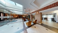 This expansive lobby makes a striking first impression through a thoughtful blend of classic and contemporary design. Exposed red brick columns and walls serve as the architectural backbone of the space, providing warmth and texture throughout. Large skylights run along the ceiling, flooding the interior with abundant natural light and creating an airy, open atmosphere that feels both grand and welcoming.

The flooring features large-format white and gray tiles arranged in a refined geometric pattern, contrasting beautifully with the richness of the brick. Elegant candelabra-style chandeliers hang throughout, complementing wall-mounted sconces that add a warm, ambient glow.

Seating areas are thoughtfully distributed across the lobby. Plush amber-toned velvet armchairs with sleek side tables create inviting conversation clusters, while a lounge nook features a pair of tufted gray Chesterfield sofas centered around a dark coffee table, offering a more relaxed setting bathed in natural light from floor-to-ceiling windows.

A reception desk anchors one section of the space, with a bike storage area visible nearby. A glass-enclosed area provides a semi-private zone within the open floor plan. A separate room features a striking marble-clad accent wall with an illuminated aquarium display set into its center, adding a unique focal point. Lush tropical plants are placed throughout, enhancing the lobby's vibrant, polished ambiance.