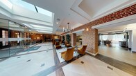 This expansive lobby makes a striking first impression through a thoughtful blend of classic and contemporary design. Exposed red brick columns and walls serve as the architectural backbone of the space, providing warmth and texture throughout. Large skylights run along the ceiling, flooding the interior with abundant natural light and creating an airy, open atmosphere that feels both grand and welcoming.

The flooring features large-format white and gray tiles arranged in a refined geometric pattern, contrasting beautifully with the richness of the brick. Elegant candelabra-style chandeliers hang throughout, complementing wall-mounted sconces that add a warm, ambient glow.

Seating areas are thoughtfully distributed across the lobby. Plush amber-toned velvet armchairs with sleek side tables create inviting conversation clusters, while a lounge nook features a pair of tufted gray Chesterfield sofas centered around a dark coffee table, offering a more relaxed setting bathed in natural light from floor-to-ceiling windows.

A reception desk anchors one section of the space, with a bike storage area visible nearby. A glass-enclosed area provides a semi-private zone within the open floor plan. A separate room features a striking marble-clad accent wall with an illuminated aquarium display set into its center, adding a unique focal point. Lush tropical plants are placed throughout, enhancing the lobby's vibrant, polished ambiance.