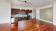 This well-appointed single-floor residence blends modern design with refined materials throughout its living spaces. The open-concept kitchen serves as a centerpiece, showcasing rich walnut cabinetry extending floor to ceiling, dark granite countertops, a large undermount stainless steel sink, and a full suite of stainless steel appliances including a built-in oven, microwave, cooktop, refrigerator, and dishwasher. A kitchen island with matching granite provides additional prep space and opens directly to the main living area, which features gleaming cherry hardwood floors and a striking architectural column. Distinctive glass block walls appear throughout the space, diffusing soft, ambient natural light while maintaining privacy — a standout design feature that lends the home a unique character. The bedroom area includes generous built-in shelving and closet storage, with neutral carpeting underfoot. Two bathrooms offer contrasting yet equally refined aesthetics: one features marble tile flooring and countertops with polished hardware, while the other offers a frameless glass-enclosed shower, wood vanity, white countertop, and subway tile accents. Together, these spaces create a cohesive, modern living environment with thoughtful attention to material quality and architectural detail.