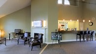 Located in Brandywine, this single-story space offers an inviting and well-appointed interior designed for comfort and community. The lounge area serves as a central gathering point, anchored by a classic white-mantled fireplace that adds a traditional, residential charm to the space. A wall-mounted flat-screen television with flanking sconce lighting is positioned above the fireplace surround, creating a functional and stylish focal point. Plush, dark leather armchairs are arranged nearby, offering a comfortable seating arrangement ideal for relaxed conversation or quiet leisure.

The soaring ceiling height lends an open, airy quality to the interior, and an arched transom window near the roofline draws in natural light, enhancing the bright, welcoming atmosphere. Neutral sage-green walls and carpeted floors in muted tones contribute to a calm, cohesive aesthetic throughout.

Adjacent to the lounge, a service counter with pendant lighting and a granite-style surface appears to function as a refreshment or reception area, further supporting the space's communal purpose. A seasonal display easel adds a personable, community-oriented touch.