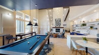Located at 15270 Voss Rd in Sugar Land, Texas, this single-story community clubhouse is a thoughtfully designed social hub that blends contemporary aesthetics with functional amenity spaces. The main gathering area commands attention with dramatically soaring ceilings, a striking chandelier, and an abundance of natural light streaming through oversized windows that frame views of the surrounding exterior. A herringbone-patterned light wood floor runs throughout, lending warmth and cohesion to the open layout.

At the heart of the space, a full-sized billiards table anchors the recreation zone, complemented by pendant lighting overhead. Adjacent to it, a comfortable lounge area centers around a floor-to-ceiling marble fireplace, surrounded by curated seating including leather and upholstered chairs, ottomans, and layered area rugs in deep blue tones. A large abstract artwork above the fireplace adds a gallery-quality focal point.

The kitchenette area is outfitted with bold navy blue cabinetry, a white quartz countertop, a marble-slab backsplash, open display shelving, and a stainless steel sink — ideal for catering and community events. A cozy dining nook with banquette seating and a marble-top table provides an intimate gathering spot. A separate lounge alcove with tufted accent chairs and high walls offers a quieter retreat within the space.