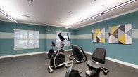 This fitness center is designed to support an active lifestyle within a compact, thoughtfully arranged space. The room features a distinctive two-tone color scheme, with bold teal walls accented by a wide white horizontal stripe that adds a sense of visual depth and modernity. Crown molding runs along the upper perimeter, lending a polished, finished look to the space.

The facility is equipped with a selection of cardio machines, including upright exercise bikes and an elliptical trainer, all positioned to allow comfortable use and easy movement between equipment. The flooring consists of durable, dark speckled fitness-grade material, well-suited for high-traffic use.

Overhead, a combination of recessed lighting and a track-style linear fixture ensures the space is brightly and evenly illuminated. Two windows fitted with horizontal blinds allow for natural light to filter in, enhancing the open feel of the room. A contemporary geometric wall art piece in yellow, white, and gray adds a decorative touch, contributing to the space's energetic yet welcoming ambiance.

The single-floor layout is practical and efficient, making it easy to navigate and use. Overall, the fitness center strikes a balance between function and aesthetic, providing a motivating environment for exercise.