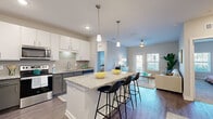 This single-floor residence is designed with a contemporary open-concept layout that seamlessly connects the kitchen, dining, and living areas. The kitchen is a standout feature, equipped with stainless steel appliances, two-tone cabinetry pairing white upper cabinets with gray lower cabinets, a tile backsplash, and granite countertops that extend across a large central island. The island accommodates bar seating for multiple guests and is illuminated by sleek pendant lights overhead, creating an inviting focal point for the space. Wood-style flooring runs throughout the main living area, complementing the neutral gray walls and recessed lighting.

The living room receives natural light through windows facing the exterior and offers a comfortable, open atmosphere. Two carpeted bedrooms each feature multiple windows with exterior views, providing ample natural light. The primary bedroom includes a walk-in closet with wire shelving. The bathroom features a tub-shower combination with tile surround, wood-style flooring, and granite countertop vanity. A dedicated in-unit laundry room houses a full-size washer and dryer with overhead shelving for added storage convenience.