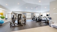 Located in Athens, Alabama, this fitness center offers a comprehensive workout environment designed to accommodate a wide range of exercise needs. The space is housed on a single level and is generously sized, providing ample room for movement and equipment placement without feeling crowded.

The room features a striking vaulted ceiling with recessed lighting and a ceiling fan, contributing to an open, airy atmosphere. Neutral-toned walls in soft gray and beige are complemented by multiple windows fitted with horizontal blinds, allowing natural light to fill the space while maintaining privacy.

Durable rubber flooring covers the workout areas, providing safety and cushioning underfoot. The cardio section is well-appointed with treadmills, elliptical machines, stair climbers, and stationary and cycling bikes, many equipped with individual monitors. A strength training area features a multi-station cable machine, free weights including a tiered dumbbell rack with a broad range of weights, and adjustable benches for varied exercises. Stability balls and additional accessories are also available.

A wall-mounted television and mirrors enhance the functionality of the space. Entry is provided through double glass doors, and a water fountain and built-in storage shelving add practical amenities. The overall design is clean, modern, and purpose-built for an effective fitness experience.