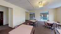 This single-floor community room is a functional, multi-purpose gathering space designed to accommodate a variety of social activities. The main gathering area is furnished with several rectangular tables and stackable chairs arranged to support group dining or meetings, all set on durable carpeted flooring. A ceiling fan with a light fixture provides overhead comfort, while large windows and glass partitions offer direct views into an adjoining indoor pool and patio area, creating a bright, connected atmosphere.

A dedicated kitchen area supports catering and event needs, featuring white cabinetry both above and below the counter, a double-basin sink, an electric range, a full-size refrigerator, and a microwave. The tile flooring and ample counter space make this kitchen practical for preparing and serving refreshments.

A secondary seating alcove provides a quieter retreat within the space, lined with white horizontal shiplap-style paneling and wall-mounted sconce lighting. A sliding window in this area frames an outdoor view, allowing natural light to filter in. Together, these distinct zones make the community room a well-rounded space suited for gatherings, events, and casual socializing.