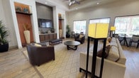 This single-floor community space is designed with a cohesive, contemporary aesthetic that balances comfort and functionality. The main lounge area serves as the centerpiece of the space, featuring soaring tray ceilings with a ceiling fan, recessed lighting, and rich hardwood-style flooring. A geometric diamond-patterned area rug anchors the seating arrangement, which includes dark leather chairs, a round coffee table, and additional lounge seating. A built-in media wall with a large flat-screen television and dark wood cabinetry adds a refined focal point, complemented by decorative niches displaying curated décor and tall vases. Large windows line one wall, flooding the interior with natural light and framing views of palm trees and an outdoor patio and pool area. A dining nook near the exit door offers an intimate setting with a small bistro-style table and direct sightlines to the covered outdoor lounge. The kitchen area features dark espresso cabinetry, granite countertops with a bar-height seating ledge, a mosaic tile backsplash, and recessed lighting throughout. A welcoming entryway with a framed mirror, wall sconces, and tasteful greenery completes the inviting atmosphere of this communal gathering space.