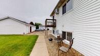 Located at 1262 W Lincoln Hwy in DeKalb, Illinois, this single-floor unit offers a practical and comfortable layout with a variety of notable exterior features. The building is clad in white vinyl siding and presents a well-maintained appearance with multiple ground-level windows that allow natural light into the interior spaces.

The unit is accessed through a private entry door with glass panel detailing, accented by a small gravel seating area just outside — ideal for relaxing outdoors. This area includes space for a small bistro table and chairs, lending a cozy outdoor retreat directly off the unit. An elevated wooden deck is also present on the building's exterior, providing an additional outdoor vantage point overlooking the surrounding grounds.

The property includes a detached garage structure with multiple bays, offering convenient off-street parking and additional storage. A concrete driveway connects the parking area to the main building.

The expansive backyard is a standout feature, offering generous open green space enclosed by a solid wood privacy fence. This fenced yard provides a sense of seclusion and ample room for outdoor activities. Lush, well-kept grass extends throughout, enhancing the overall appeal of the outdoor environment. The combination of private access, outdoor amenities, and generous yard space makes this property a well-rounded option in the DeKalb area.