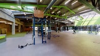 Pickwick Plaza Gym in Kansas City, Missouri offers a expansive, open-plan fitness environment housed within a repurposed industrial structure. The architectural character is defined by soaring exposed steel trusses painted in vibrant lime green, visible ductwork, and original brick walls that preserve the building's historic character. Skylights and large clerestory windows flood the interior with natural light, enhancing the airy, warehouse-style atmosphere.

The main training floor is finished with durable rubber flooring and organized into distinct functional zones. A strength training area features a multi-station power rack system with pull-up attachments, free weights, kettlebells, and weight plates neatly organized along mirrored walls. A dedicated cardio section lines one brick wall, equipped with treadmills, elliptical machines, and stair climbers, each paired with individual monitors. The open functional training zone includes TRX suspension trainers, battle ropes, and hanging heavy bags suspended from the overhead structure. A separate, quieter area near the entrance provides water stations and additional equipment storage. Bold green-and-black motivational signage adds an energetic, athletic ambiance throughout the facility.