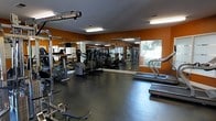 This fitness center is a dedicated single-floor workout facility designed to support a comprehensive exercise routine. The space is finished with durable dark rubber flooring, providing a safe and stable surface for a variety of workouts. Bold orange accent walls lend an energetic ambiance that complements the active nature of the space, while large wall-mounted mirrors span the length of the room, creating a sense of expanded space and allowing users to monitor their form during exercise. Natural light filters in through a window overlooking outdoor greenery, contributing to a bright and motivating environment supplemented by overhead fluorescent lighting. The fitness center is outfitted with an impressive array of equipment, including treadmills, elliptical machines, and a variety of strength-training cable machines and multi-use workout stations. Equipment is thoughtfully arranged to allow for comfortable movement throughout the room. The layout balances cardio and strength zones effectively, making the space functional for users with varying fitness goals. Overall, this fitness center offers a well-rounded, practical, and inviting environment for daily exercise.