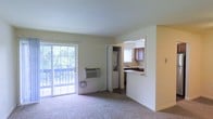 Located at 400 W Crestwood Dr in Camp Hill, Pennsylvania, this single-floor residence offers a practical and comfortable open-concept layout. The living area is carpeted and bathed in natural light, thanks to a large sliding glass door that leads directly to a private outdoor balcony overlooking lush greenery — a standout feature that brings the outdoors in. A wall-mounted HVAC unit ensures year-round climate control within the living space. Just off the living area, a conveniently positioned closet houses a stacked in-unit washer and dryer, adding a layer of everyday practicality. The kitchen is accessed through an open pass-through cutout, creating a connected feel between the living and cooking spaces. It features rich medium-toned cabinetry, a classic tile backsplash, dark countertops, and a window that allows additional natural light to filter in. The warm yellow accent wall adds a touch of character to the space. Overall, the unit presents a functional and bright single-level layout well-suited for comfortable everyday living in the Camp Hill area.