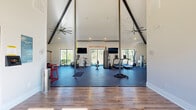 Located in Athens, Georgia, this fitness center offers a generously sized, single-floor exercise space designed to support a wide range of workout routines. The facility is immediately distinguished by its dramatic vaulted ceiling with exposed dark steel beam trusses, creating an airy, open atmosphere that feels expansive and energizing. Large industrial ceiling fans are mounted high above, ensuring consistent airflow throughout the space.

The main exercise area features durable rubber flooring and is equipped with an impressive array of cardio machines, including treadmills, ellipticals, a stationary bike, and a rowing machine. Strength training is well-represented with a power rack, free weights, battle ropes, a heavy punching bag, a TRX suspension system, plyo boxes, and medicine balls. A large wall mirror in the training zone enhances spatial perception and supports form-focused workouts.

Natural light filters in through multiple large windows along the perimeter, offering views of the surrounding outdoor environment. Wall-mounted televisions provide entertainment during workouts, while bold motivational phrases — including "Hustle for Muscle" and "Big Results" — are displayed prominently on the walls. The entry foyer features hardwood-style flooring, built-in storage cubbies, yoga mat storage, and double glass doors, creating a welcoming and organized arrival experience.
