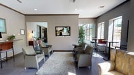 This well-appointed single-floor clubhouse offers a thoughtfully designed gathering space with distinct zones for relaxation and socializing. The main lounge area features patterned upholstered armchairs arranged around a plush shag area rug, a wall-mounted flat-screen television, and dark wood crown molding that frames the neutral-toned walls. High bar-height tables and stools positioned near large windows provide a casual seating alternative with ample natural light.

A standout feature of the space is the bright solarium-style lounge, distinguished by a vaulted teal-painted ceiling with a ceiling fan, floor-to-ceiling windows, and direct views of an outdoor pool area. This room is furnished with grey sofas, a round wood coffee table, and a bold geometric area rug in teal, navy, and white tones. A mosaic-tiled fireplace with an eye-catching teal tile surround serves as an architectural focal point, adding warmth and visual interest.

A secondary seating alcove with large multi-pane windows offers a quieter retreat, bathed in natural light with a mix of upholstered chairs. The kitchenette is equipped with granite countertops, a teal mosaic tile backsplash, stainless steel appliances, a double sink, and ample cabinetry — ideal for event preparation. Dark tile flooring flows consistently throughout, grounding the cohesive teal and grey design palette.