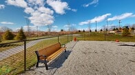 Located at 9 Monticello Dr in Brewster Village, NY, this outdoor dog park is a well-appointed amenity set within a larger residential community. The space features a generously sized, gravel-surfaced enclosure fully enclosed by black chain-link fencing, providing a safe and open area for pets to roam freely. Multiple wooden benches with black metal frames are positioned throughout the park, offering comfortable seating for owners while they supervise their animals. A classic red fire hydrant serves as a charming focal point within the enclosure. The park benefits from abundant natural light and open sky views, creating a bright and airy atmosphere. Neatly trimmed evergreen trees and maintained green lawns frame the perimeter, contributing to a pleasant outdoor setting. A pathway leads through the surrounding grounds, connecting the dog park to the broader community. The residential buildings of the community — featuring multi-story construction with neutral siding, private balconies, and large windows — provide an attractive backdrop. A colorful playground is also visible nearby, highlighting the family-friendly nature of the overall community. The space is thoughtfully designed to serve both pets and their owners in a comfortable, open-air environment.