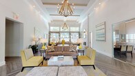 This elegant single-floor space makes an immediate impression upon entry, where a console table with a decorative mirror welcomes visitors beneath soaring ceilings that flood the foyer with natural light. The main lounge area is defined by dramatic coffered ceilings adorned with an eye-catching geometric brass chandelier, whose warm glow complements the recessed lighting throughout. Large windows along the front wall frame lush outdoor views while bathing the interior in abundant natural light.

The herringbone-patterned hardwood flooring extends seamlessly throughout the space, adding a refined architectural detail underfoot. The lounge is furnished with a carefully curated mix of plush taupe sofas, bold yellow accent chairs, patterned ottomans, and chic side tables, all tied together by a yellow-toned area rug. Accent lamps, framed artwork, and patterned throw pillows further elevate the design aesthetic.

Toward the rear, full-height glass sliding doors open to an adjacent room, visible as a dining or meeting area, offering flexible use of the space. The overall ambiance is bright, polished, and welcoming, balancing contemporary sophistication with approachable warmth.