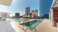 This exceptional rooftop amenity deck offers an elevated urban retreat with breathtaking, unobstructed views of a dynamic downtown skyline. The centerpiece of the space is a generously sized, crystal-clear swimming pool with a sleek, modern design, complemented by a separate spa pool with brass-finished entry rails — both finished in light stone tile that enhances the open, airy atmosphere. Surrounding the pools, a spacious sun deck is appointed with an array of loungers and stylish seating areas designed for relaxation. A standout feature is the collection of private cabanas, each outfitted with slatted wood paneling, sheer curtains, ceiling fans, and plush navy-blue cushioned seating, offering both shade and an intimate lounge experience. Umbrella-shaded seating areas provide additional options for guests seeking relief from the sun. A sleek, modern building facade with floor-to-ceiling glass panels serves as a backdrop on one side of the deck, while a glass-railed terrace along the perimeter frames sweeping views of surrounding high-rises, historic architecture, and the broader cityscape. Built-in planter beds with ornamental grasses add a touch of greenery to the elevated outdoor setting, creating a sophisticated balance of nature and urban design.