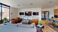 This single-floor space showcases a thoughtfully designed open-plan layout that seamlessly integrates living, dining, and kitchen areas. Reclaimed hardwood flooring runs throughout, adding warmth and texture that contrasts beautifully with the clean white walls and contemporary finishes.

The living area is anchored by a generous seating arrangement featuring a grey sofa and rich cognac leather armchairs, oriented toward a wall-mounted media console displaying multiple flat-screen televisions. Recessed lighting and in-ceiling speakers enhance the entertainment-ready atmosphere.

Large, black-framed floor-to-ceiling windows flood the interior with abundant natural light while framing striking urban views of the surrounding cityscape. A glass-paneled door opens directly onto a private outdoor terrace, extending the living space and providing an elevated outdoor retreat.

The kitchen is defined by warm natural wood cabinetry, dark countertops, stainless steel appliances, and a glossy grey tile backsplash. A substantial solid wood dining table with upholstered chairs serves as the central gathering point, bridging the kitchen and living zones.

A secondary dining area features a dark table set against expansive windows, offering additional flexibility for meetings or dining. Throughout, the space balances urban sophistication with approachable warmth.