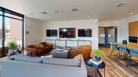 This single-floor space showcases a thoughtfully designed open-plan layout that seamlessly integrates living, dining, and kitchen areas. Reclaimed hardwood flooring runs throughout, adding warmth and texture that contrasts beautifully with the clean white walls and contemporary finishes.

The living area is anchored by a generous seating arrangement featuring a grey sofa and rich cognac leather armchairs, oriented toward a wall-mounted media console displaying multiple flat-screen televisions. Recessed lighting and in-ceiling speakers enhance the entertainment-ready atmosphere.

Large, black-framed floor-to-ceiling windows flood the interior with abundant natural light while framing striking urban views of the surrounding cityscape. A glass-paneled door opens directly onto a private outdoor terrace, extending the living space and providing an elevated outdoor retreat.

The kitchen is defined by warm natural wood cabinetry, dark countertops, stainless steel appliances, and a glossy grey tile backsplash. A substantial solid wood dining table with upholstered chairs serves as the central gathering point, bridging the kitchen and living zones.

A secondary dining area features a dark table set against expansive windows, offering additional flexibility for meetings or dining. Throughout, the space balances urban sophistication with approachable warmth.