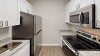 This single-floor unit offers a clean, contemporary layout designed for functional everyday living. The kitchen is well-appointed with white shaker-style cabinetry, granite-look countertops, and a full suite of stainless steel appliances, including a refrigerator, electric range, and over-the-range microwave. Durable wood-look vinyl flooring runs throughout, providing a cohesive and easy-to-maintain finish across the space. The open living area is bright and spacious, with neutral white walls and a seamless sightline into the kitchen and laundry area, where an in-unit washer is conveniently tucked beneath the cabinetry. Two well-proportioned bedrooms each feature their own closet storage — one with a reach-in closet with a hanging rod, and the other with a double-door sliding closet — both rooms continuing the dark-toned vinyl flooring for a grounded, modern feel. The bathroom is finished with large-format gray stone-look wall tile surrounding a soaking tub and shower combo, complemented by brushed nickel fixtures and granite-accented vanity details. The overall aesthetic is fresh, neutral, and move-in ready, with consistent finishes that lend the space a cohesive and updated character throughout.