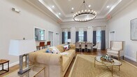 This single-floor amenity space combines classic architectural detailing with a warm, inviting atmosphere. The entryway greets visitors with large-format tile flooring, crisp white walls, crown molding, and an arched wood-and-glass front door that floods the corridor with natural light. Framed artwork and decorative greenery add a polished touch to the welcoming foyer.

The main gathering area is the centerpiece of the space, showcasing soaring tray ceilings adorned with a grand circular chandelier that casts a soft, ambient glow. Rich hardwood-style flooring flows throughout, anchoring a generously sized lounge furnished with a tufted camel-colored sofa, woven area rug, and gold-accented glass side tables. A flat-screen television is mounted on the wall nearby, enhancing the comfort and functionality of the space.

Adjacent to the lounge, a formal dining arrangement seats a large group beneath the same elegant chandelier, with upholstered chairs and tall windows dressed in two-toned navy and neutral drape panels that allow natural light to filter in gracefully. Built-in workstations with computer setups are neatly integrated into the perimeter of the room through arched passageways, offering a dedicated workspace. A service counter with bar-height seating rounds out the multifunctional layout of this thoughtfully designed community space.