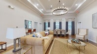 This single-floor amenity space combines classic architectural detailing with a warm, inviting atmosphere. The entryway greets visitors with large-format tile flooring, crisp white walls, crown molding, and an arched wood-and-glass front door that floods the corridor with natural light. Framed artwork and decorative greenery add a polished touch to the welcoming foyer.

The main gathering area is the centerpiece of the space, showcasing soaring tray ceilings adorned with a grand circular chandelier that casts a soft, ambient glow. Rich hardwood-style flooring flows throughout, anchoring a generously sized lounge furnished with a tufted camel-colored sofa, woven area rug, and gold-accented glass side tables. A flat-screen television is mounted on the wall nearby, enhancing the comfort and functionality of the space.

Adjacent to the lounge, a formal dining arrangement seats a large group beneath the same elegant chandelier, with upholstered chairs and tall windows dressed in two-toned navy and neutral drape panels that allow natural light to filter in gracefully. Built-in workstations with computer setups are neatly integrated into the perimeter of the room through arched passageways, offering a dedicated workspace. A service counter with bar-height seating rounds out the multifunctional layout of this thoughtfully designed community space.