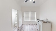 This compact studio unit offers a thoughtfully organized single-floor layout encompassing a main living area, a kitchenette, and a private bathroom. The interior is finished with clean white walls and light wood-style laminate flooring throughout, creating a bright and cohesive aesthetic.

The main living space is open and airy, benefiting from a large double-panel window fitted with white horizontal blinds that allow ample natural light to filter in. A ceiling fan with an integrated light fixture is centrally mounted, providing both ventilation and ambient lighting. The room accommodates a compact kitchenette equipped with a two-burner cooktop, an under-counter oven, a combined sink and countertop unit, and a full-height stainless steel refrigerator positioned nearby. A wall-mounted panel houses electrical controls, and a wall unit provides climate control.

The entry area features a solid white door with dual locks, maintaining security and privacy. Adjacent to the main space, the bathroom is accessible through an interior door and includes a toilet, a dedicated walk-in shower stall with a chrome showerhead and curtain rod, and an additional window for natural light and ventilation. The overall design is minimal and functional, making efficient use of every available area.