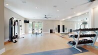 Located at Tapestry Park in Indian Land, SC, this expansive fitness center offers a comprehensive workout facility across a single, generously sized floor. The space is divided into distinct functional zones, each thoughtfully designed to support a variety of exercise modalities. Light wood-look flooring with contrasting blue accent strips runs throughout, lending a clean, modern aesthetic to the entire facility.

The main workout area features strength training equipment including cable machines, weight benches, and a full rack of dumbbells, all positioned beneath recessed lighting and ceiling fans that ensure comfort during exercise. Floor-to-ceiling mirrors line multiple walls, amplifying the sense of space and supporting proper form during workouts.

A dedicated cardio zone houses elliptical trainers and a stair climber, positioned near large windows and glass doors that invite natural light and offer views of the surrounding greenery. A separate flex studio area, accented by distinctive vertical-stripe wall paneling and a stylized ceiling detail, provides room for stretching, group exercise, and functional training with stability balls and resistance equipment.

Wall-mounted televisions are installed throughout, and ceiling fans maintain airflow across all zones. The facility is accessible via multiple entry points, with glass-paneled French doors connecting sections and leading to outdoor areas, creating a bright, open, and inviting environment for all fitness levels.