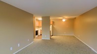 This single-floor residential unit offers a practical and functional layout designed for comfortable everyday living. The main living area is spacious and open, finished with wall-to-wall carpet flooring in a neutral beige tone that complements the warm, tan-painted walls throughout. A ceiling fan is centrally mounted, providing both lighting and airflow to the living space. The open floor plan connects seamlessly to the kitchen, which is partially separated by a structural column, adding subtle architectural definition between the two areas. The kitchen is well-equipped, featuring light wood cabinetry, white appliances including a refrigerator, and a dark-finish range, all set against a clean, light-colored countertop and smooth flooring. White baseboards run along the perimeter of the living area, providing a crisp contrast to the carpeted floors and neutral walls. The overall ambiance is calm and understated, with a cohesive color palette of beige and warm neutrals creating a welcoming atmosphere. The open connectivity between the living and kitchen areas makes the space feel airy and versatile, well-suited for a variety of furnishing arrangements and everyday use.