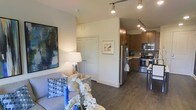 This single-floor residence features an open-concept layout that seamlessly connects the living area and kitchen, creating a cohesive and functional living environment. The living area is anchored by a comfortable gray sofa accented with blue decorative pillows, complemented by a sleek table lamp and bold abstract wall art that adds a pop of color and personality to the space. Dark hardwood-style flooring runs throughout, providing a warm and grounding contrast to the neutral, light-toned walls.

The kitchen is well-appointed with warm-toned wooden cabinetry, stainless steel appliances including a refrigerator, range, and microwave, and a practical layout designed to maximize efficiency. Track lighting installed along the ceiling illuminates both the kitchen and the adjoining living area, ensuring ample light across the open floor plan.

A set of white panel doors leads to what appears to be a closet or utility space, neatly integrated into the layout. Framed artwork on the walls adds thoughtful decorative touches throughout the space. Overall, the residence offers a clean, modern aesthetic with a functional and well-organized design.