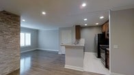 This single-floor space showcases a thoughtfully designed open-concept layout that seamlessly connects the kitchen and living area. The kitchen is a standout feature, equipped with stainless steel appliances, including a refrigerator and gas range, complemented by warm medium-toned wood cabinetry. A peninsula with a light granite countertop and an integrated sink serves as a functional divider between the kitchen and the adjacent living space, while a classic herringbone tile backsplash adds a refined decorative touch. The kitchen area is finished with light-toned tile flooring, which transitions smoothly into the rich, medium-toned hardwood-style flooring that extends throughout the open living area. Recessed lighting is installed throughout the ceiling, providing bright, even illumination across the entire space. The walls are finished in a neutral gray tone, creating a clean and contemporary ambiance. White baseboards and trim offer crisp contrast against the gray walls. A window in the living area allows natural light to filter in, enhancing the airy, open feel of the space.