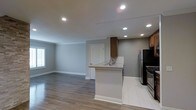 This single-floor space showcases a thoughtfully designed open-concept layout that seamlessly connects the kitchen and living area. The kitchen is a standout feature, equipped with stainless steel appliances, including a refrigerator and gas range, complemented by warm medium-toned wood cabinetry. A peninsula with a light granite countertop and an integrated sink serves as a functional divider between the kitchen and the adjacent living space, while a classic herringbone tile backsplash adds a refined decorative touch. The kitchen area is finished with light-toned tile flooring, which transitions smoothly into the rich, medium-toned hardwood-style flooring that extends throughout the open living area. Recessed lighting is installed throughout the ceiling, providing bright, even illumination across the entire space. The walls are finished in a neutral gray tone, creating a clean and contemporary ambiance. White baseboards and trim offer crisp contrast against the gray walls. A window in the living area allows natural light to filter in, enhancing the airy, open feel of the space.