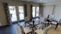 This single-story Dallas, TX home combines contemporary design with comfortable living. The dining area serves as a focal point of the space, showcasing dark hardwood flooring that contrasts beautifully with the neutral, warm-toned walls. A round glass-top dining table surrounded by white upholstered chairs with dark wood frames anchors the room, creating an inviting gathering space. Two sets of white French doors flank the dining area, each dressed with bold black-and-white geometric patterned curtains that add a striking decorative element. These doors flood the interior with natural light while providing seamless access to the outdoor patio area, visible beyond. Recessed lighting in the ceiling complements the natural light, ensuring the space is well-lit at any time of day. A neutral area rug grounds the dining arrangement, adding texture and warmth to the dark floors. The overall ambiance is one of modern elegance — clean lines, a cohesive monochromatic palette punctuated by bold accents, and a strong indoor-outdoor connection that is well-suited to the Dallas lifestyle.