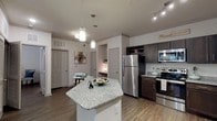 This single-floor residence offers a thoughtfully designed layout that balances functionality with contemporary style. The kitchen serves as a central highlight, showcasing rich dark cabinetry paired with granite countertops, a suite of stainless steel appliances including a refrigerator, range, and microwave, and a striking hexagonal island with pendant lighting overhead. The hardwood-style flooring runs throughout, adding warmth and cohesion to the space. Track lighting complements the pendant fixtures, ensuring the kitchen and adjoining areas are well illuminated. The open layout connects the kitchen to a comfortable living area, which features large windows dressed with sheer curtains and blinds, allowing natural light to fill the room. The bedroom is a serene retreat, appointed with a ceiling fan, generous windows with patterned drapes, and ample space for furnishings. The bathroom continues the design language established in the kitchen, featuring dark wood vanity cabinetry with granite countertops, a single basin sink, and a full tub-shower combination. Clean white walls and hardwood-style flooring carry through this room as well, maintaining a consistent and polished aesthetic throughout the entire residence.