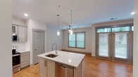 This single-floor residence offers a well-designed open-concept layout that seamlessly connects the kitchen, dining, and living areas. The kitchen serves as the focal point, anchored by a large central island with a quartz countertop, an undermount sink, and pendant lighting above. Grey shaker-style cabinetry lines the perimeter walls, complemented by a white tile backsplash and a full suite of stainless steel appliances, including a refrigerator, electric range, built-in microwave, and dishwasher. Light hardwood flooring runs throughout the kitchen and living areas, enhancing the bright, airy atmosphere created by multiple windows fitted with horizontal blinds. The living area benefits from a ceiling fan and direct outdoor access through a set of glass-paneled French doors topped with transom windows, allowing an abundance of natural light. The bedroom is carpeted for comfort, featuring its own ceiling fan and a window with outdoor views. The bathroom is finished with wood-look tile flooring, a grey vanity with a white countertop, a wall-mounted mirror, overhead cabinetry, and a walk-in shower with glass enclosure.