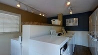 This single-floor laundromat offers a practical and organized environment designed with customer convenience in mind. The space features a row of top-loading washing machines and dryers neatly arranged along a central divider, providing an efficient workflow for patrons. White paneling lines the lower half of the divider walls, lending a clean and tidy aesthetic throughout the facility. The upper walls are painted in contrasting bold tones — a warm golden-ochre on one side and a deep navy blue on the other — giving the space a distinct visual character. Overhead, track lighting runs along the ceiling, directing bright, focused light across the work surfaces and machine areas to ensure clear visibility. The textured ceiling adds a subtle architectural detail to the otherwise straightforward interior. Instructional signage is posted on the walls, providing helpful guidance for operating the machines. A laundry basket is stored on a shelf above the divider, and the overall layout reflects a well-maintained facility with an emphasis on functionality and ease of use. The space is designed to accommodate multiple users simultaneously, making it a practical solution for laundry needs.