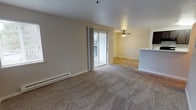 Located at 1206 NW Tahoe Lane in Silverdale, Washington, this single-floor residence offers a comfortable and functional layout designed for everyday living. The main living area is generously sized, finished with wall-to-wall carpeting that adds warmth underfoot, and features a ceiling fan for year-round comfort. A baseboard heating system runs along the exterior wall, ensuring efficient climate control throughout the space. Neutral, light-toned walls create a bright and airy atmosphere, complementing the natural light that filters in through a sliding glass door. This door opens directly onto a private balcony, providing a seamless connection to the outdoors and views of the surrounding neighborhood. The transition from the carpeted living area leads into a secondary space with wood-look flooring, adding a subtle design contrast between the zones. A partial kitchen counter with bar-style seating is visible at the edge of the living space, suggesting an open-concept layout that keeps the kitchen and living areas connected. The overall ambiance is clean, understated, and move-in ready, making it a practical and welcoming home in the Silverdale community.