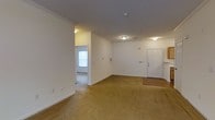 This single-floor space offers a straightforward and functional layout centered around a generously sized open living area. The room is finished with wall-to-wall beige carpeting and crisp white walls, creating a clean, neutral canvas throughout. Crown molding lines the ceiling, adding a subtle architectural detail that elevates the overall finish of the space. Ceiling-mounted light fixtures provide consistent ambient lighting across the room.

A paneled front entry door is positioned along one wall, transitioning from a small hardwood-floored entry area into the carpeted main living space. Beside the entry, a utility panel is recessed into the wall, keeping functional elements neatly integrated. The textured ceiling adds a classic residential character throughout.

An open passageway leads to an adjoining room, visible from the main living area, which benefits from a window that introduces natural light into the interior. This secondary space mirrors the carpeted flooring and white wall treatment of the main area, maintaining a cohesive aesthetic throughout the unit. Together, the rooms provide a versatile, open feel well-suited for a variety of living arrangements.