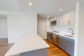This well-appointed one-bedroom, one-bathroom residence in Cambridge, MA offers a thoughtfully designed single-floor layout with clean, modern finishes throughout. The open-concept living and dining area is bathed in natural light from large floor-to-ceiling windows, complemented by warm hardwood flooring that flows seamlessly through the main living spaces. The contemporary kitchen is a standout feature, boasting a central island with white quartz countertops, two-tone cabinetry in grey and white, and a full suite of stainless steel appliances including a refrigerator, range, microwave, and dishwasher. Recessed lighting illuminates the space efficiently. A hallway connects the living area to the private bedroom, which features plush carpeting and oversized windows offering expansive city views of the Cambridge and Boston skyline. Two closets provide ample storage throughout the unit. The bathroom is finished with a tiled tub-shower combination, a vanity with under-cabinet storage, and hardwood-style flooring. An in-unit laundry area adds everyday convenience. The neutral white walls and clean architectural lines create a bright, modern ambiance throughout this refined urban residence.