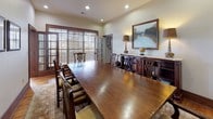 This single-room space exudes traditional elegance, centered around a substantial solid hardwood dining or conference table surrounded by a set of Chippendale-style chairs with upholstered seats. The warm-toned hardwood flooring runs throughout and is partially covered by a decorative area rug, adding texture and a sense of refinement to the space.

Recessed lighting set into the smooth ceiling provides even, functional illumination, while a pair of classic table lamps atop a richly detailed wooden sideboard along one wall contribute a warm, ambient glow. The sideboard itself is a notable architectural accent, featuring carved detailing consistent with the room's traditional aesthetic. Above it hangs a landscape oil painting in a dark wood frame, complementing the nature-themed framed botanical prints displayed on the adjacent wall.

Large French doors fitted with wooden blinds allow natural light to filter in while maintaining privacy, and a secondary doorway opens to a connecting hallway or adjacent sitting area, suggesting this space is part of a larger facility. The neutral, soft white walls keep the focus on the rich woodwork and furnishings. Overall, the room presents a polished, formal atmosphere well-suited for private meetings, intimate dining, or gatherings requiring a sophisticated setting.
