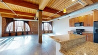 This 2-bedroom, 2-bathroom loft showcases a striking industrial aesthetic, combining raw architectural elements with functional living spaces across a single floor. Exposed wood beam ceilings, polished concrete floors, and visible ductwork define the character of the space, while structural steel and concrete columns add to its urban, warehouse-inspired charm. Accent brick walls and warm wood tones provide visual contrast throughout.

The open-plan living, dining, and kitchen area forms the heart of the home, offering a generous, light-filled expanse. The kitchen is equipped with warm-toned wood cabinetry, granite-style countertops, black appliances including a range, microwave, dishwasher, and a side-by-side refrigerator with ice dispenser. An in-unit laundry area is conveniently accessible nearby.

Both bedrooms continue the industrial aesthetic with polished concrete floors, exposed beam ceilings, and ceiling fans. The spacious primary bedroom includes an en-suite bathroom featuring a curved glass-block shower enclosure. The second bathroom offers a sleek, modern design with a glass-top vanity, open shelving, and a walk-in shower. Two closets provide ample storage. The overall ambiance is bold and contemporary, ideal for those who appreciate loft-style living.