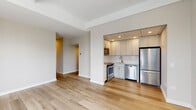 This single-floor residence features a clean, modern design highlighted by light hardwood flooring that flows throughout the main living areas. The open-concept layout connects a generously sized living area to a semi-enclosed kitchen, separated by a structural column that defines each space while maintaining visual connectivity.

The kitchen is well-appointed with white shaker-style cabinetry extending to the ceiling, light quartz countertops, a white subway tile backsplash, and a full suite of stainless steel appliances including a range, over-the-range microwave, dishwasher, and refrigerator. A recessed drop ceiling with integrated lighting adds definition to the kitchen zone.

The bedroom features light hardwood flooring, neutral walls, and a window that allows natural light to filter in. It is complemented by a spacious walk-in closet outfitted with wire shelving and carpeted flooring for added comfort underfoot.

Two bathrooms serve the unit. One includes a combined tub and shower with a sliding glass enclosure, a single-sink vanity with white cabinetry, and tile flooring. The second bathroom features a standalone glass-enclosed shower, a white vanity, and matching tile flooring. Neutral tones and clean finishes create a consistent, cohesive aesthetic throughout.