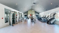 Located at 1010 Rosefinch Landing in Hoschton, GA, this single-story fitness center is a well-appointed community amenity designed to support a full spectrum of workout routines. The space is defined by its striking vaulted ceilings, which soar dramatically overhead and are fitted with recessed lighting and ceiling fans, creating an open, airy atmosphere throughout. Light-toned herringbone-patterned flooring runs the length of the facility, lending a clean and modern aesthetic underfoot.

The main floor is organized into distinct workout zones. The cardio area lines one side of the room with a robust selection of treadmills, elliptical trainers, stair climbers, stationary bikes, and a rowing machine, all positioned alongside large windows that invite natural light and views of the surrounding community. Opposite, a strength training zone houses a variety of cable and weight machines, benches, and free-weight stations. A dedicated free-weight and functional training alcove features floor-to-ceiling mirrors, a flat-screen television, a dumbbell rack, adjustable benches, a punching bag, and a well-stocked rack of kettlebells and medicine balls. Additional selectorized machines and a multi-functional cable system round out the equipment offerings, ensuring a comprehensive training experience for users of all fitness levels.