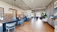 This single-story community clubhouse offers a thoughtfully designed, multi-functional layout spanning a generously sized footprint across six distinct areas. The entrance greets visitors with a polished reception desk finished in deep navy, accented by decorative pendant lighting and a herringbone wood-paneled feature wall — setting an immediately professional yet welcoming tone. Wide-plank wood-look flooring flows seamlessly throughout, tying each space together cohesively.

The main lounge area is anchored by soaring vaulted ceilings, oversized windows that flood the room with natural light, and a built-in media wall with navy-accented shelving. Comfortable seating arrangements make it an ideal gathering space. Adjacent corridors feature bold blue accent walls, textured wallcovering, and lush tropical plants, creating a vibrant, resort-inspired atmosphere.

The kitchen and dining area is highlighted by a large white waterfall island with bar seating, geometric pendant lights, dark navy cabinetry, and a striking painted brick accent wall. A connected outdoor patio area is visible through expansive windows.

The fitness center is well-appointed with strength and cardio equipment, floor-to-ceiling mirrors, a teal tray ceiling detail, and durable rubber flooring — providing a fully functional workout environment with ample natural light from surrounding windows.