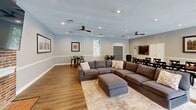 Located at 7880 Chatwell Drive in St. Louis, Missouri, this single-floor community clubhouse offers a generously sized, open-concept living and dining space designed for gathering and relaxation. The interior is anchored by a striking floor-to-ceiling exposed brick fireplace, which serves as a dramatic focal point and adds warmth and character to the room. A large flat-screen television is mounted above the fireplace, making the space ideal for communal entertainment.

The living area features a large sectional sofa and ottoman arranged on a neutral area rug, all set atop rich hardwood flooring that runs throughout the space. Dual ceiling fans with recessed lighting provide comfort and illumination across the open layout. Classic wainscoting lines the lower walls, complementing the soft, neutral gray paint above.

The dining area includes multiple high-top tables with industrial-style metal stools, positioned near a wall of large plantation-shuttered windows and a glass-paned door that opens to views of an outdoor pool. Natural light flows generously into the space, creating a bright and inviting atmosphere. The entrance features an elegant arched doorway framed by double gray panel doors, and a branded welcome mat adds a community identity touch. A kitchenette is also visible in the background, rounding out this versatile, well-appointed gathering space.