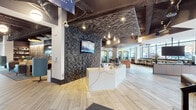 Welcome to Anova at Union Square, a thoughtfully designed single-floor commercial space that masterfully blends industrial aesthetics with modern comfort. The entrance greets visitors with a striking reception area showcasing a sculptural white marble-patterned desk set against a dramatic textured stone accent wall. Overhead, an exposed ceiling with raw beams and decorative wood-paneled sections adds industrial character, while pendant lighting creates a warm, inviting atmosphere.

Beyond the reception, the space opens into a sophisticated lounge featuring built-in wood shelving, a marble-surround gas fireplace, a wall-mounted television, and plush navy blue seating arranged around an artisan coffee table — all crowned by a bold gold sputnik pendant light. Adjacent to the lounge, an intimate banquette nook offers a semi-private setting for focused work or small meetings.

A large open area serves as a recreation and social hub, complete with a ping pong table, foosball, bar-height seating, and vibrant artwork against exposed brick walls. Large floor-to-ceiling windows flood the space with natural light and frame street-level views of the surrounding neighborhood.

Toward the rear, a versatile event or flex space with light hardwood floors and expansive glass doors opens directly onto an outdoor terrace, seamlessly connecting the interior to alfresco seating and gathering areas.