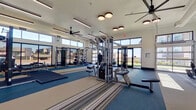This expansive fitness center offers a comprehensive workout environment designed to accommodate a wide range of exercise needs. The single-floor layout is generously proportioned, providing ample space for members to move freely between equipment zones without feeling crowded.

The facility is bathed in natural light, courtesy of large floor-to-ceiling windows and transom windows positioned along multiple walls, offering views of the surrounding exterior. Large-blade ceiling fans are distributed throughout, promoting air circulation and comfort during workouts.

The cardio zone is well-stocked with treadmills, ellipticals, stationary bikes, and a rowing machine, all arranged along windowed walls to enhance the exercise experience. The strength training area features a multi-station cable machine system, a dedicated power rack with weight plates and barbells mounted on wall-mounted storage, and a robust dumbbell rack offering a full range of free weights.

Flooring transitions between rubberized mat sections and striped carpet tiles, clearly delineating workout zones. Full-length mirrors line key walls, amplifying the sense of space and allowing users to monitor their form. Motivational neon signage adds a modern, energetic ambiance. The overall design is clean and contemporary, with white walls, structured ceiling beams, and a bright, welcoming atmosphere throughout.