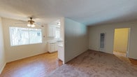 This single-floor residential unit offers a practical and functional layout centered around an open-concept living and kitchen area. The living space is finished with wall-to-wall carpet, providing a comfortable feel, while the kitchen area transitions to hardwood-style flooring. A textured popcorn ceiling runs throughout, and a ceiling fan with a light fixture in the living area ensures air circulation and ambient lighting. Natural light filters into the space through windows positioned near the kitchen, brightening the interior. The kitchen features white cabinetry, a countertop with a built-in sink, and a clean, neutral color palette. A half-wall partition separates the kitchen from the living area, maintaining an open flow while defining each zone. An additional carpeted room is visible, offering a versatile space that can serve various purposes. A wall-mounted heating or cooling unit is installed in the secondary room, providing climate control. The walls throughout are finished in a light, neutral tone, contributing to a bright and airy atmosphere. Overall, the unit presents a straightforward, well-organized layout suited for everyday living.