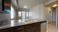 This single-floor residence is designed with an open-concept layout that seamlessly connects the kitchen and living areas, creating a functional and cohesive living space. The kitchen is well-appointed with dark espresso-toned cabinetry reaching toward the elevated ceilings, stainless steel appliances including a refrigerator, range, and microwave, and a peninsula with a double stainless steel sink and laminate countertops. The peninsula serves as a natural divider between the kitchen and the adjoining living area, which features soft carpeting and multiple windows dressed with vertical blinds that allow ample natural light to filter in. A door from the living area provides access to an outdoor space. Light wood-style flooring runs through the kitchen and bathroom areas, offering a clean contrast to the carpeted rooms. The bathroom includes a wide vanity with a single basin sink, generous countertop space, and a large mirror. Additional bedrooms are carpeted and well-proportioned, each featuring windows that bring in natural light. The overall palette of warm beige and neutral tones throughout the space contributes to a calm, move-in ready ambiance.